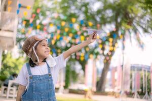 A young girl wearing a pilot's hat and a white shirt is holding a paper airplane photo