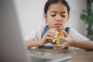 un joven niña es sentado a un mesa con un de rubik cubo en su mano foto