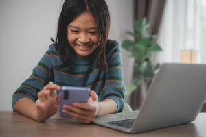 un joven niña es sentado a un mesa con un ordenador portátil y un célula teléfono. ella es mirando a el célula teléfono y sonriente foto