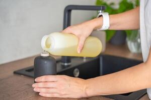 un mujer vierte jabón o detergente desde reciclado embalaje dentro un reutilizable botella en cocina. Respetuoso del medio ambiente estilo de vida concepto foto