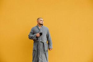 Portrait of a young short-cut bearded man in a bathrobe holding a cup of hot coffee or tea on a yellow background. The concept of morning coffee, morning, evening coffee photo