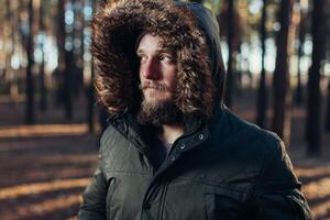 Portrait, close-up of young stylishly serious man with a beard dressed in rgreen winter jacket with a hood and fur on his head stands against the background of pine forest. photo