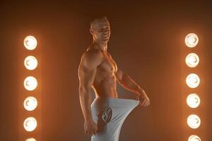 Great male size. Young muscular guy pulling back his pants and showing thumb up gesture, being proud with his genitals, lamps illumination on background photo