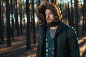 Portrait, close-up of young stylishly serious man with a beard dressed in rgreen winter jacket with a hood and fur on his head stands against the background of pine forest. photo