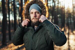 Portrait, close-up of young stylishly serious man with a beard dressed in rgreen winter jacket with a hood and fur on his head stands against the background of pine forest. photo