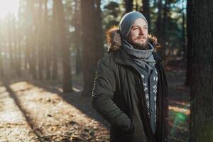 Portrait, close-up of young stylishly serious man with a beard dressed in rgreen winter jacket with a hood and fur on his head stands against the background of pine forest. photo