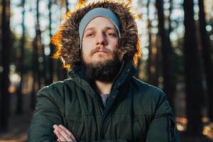 Portrait, close-up of young stylishly serious man with a beard dressed in rgreen winter jacket with a hood and fur on his head stands against the background of pine forest. photo