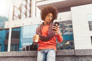 un hermoso pelo rizado chico de mezclado carrera es vestido en un de moda estilo juvenil bufanda y suéter escribe un mensaje en un telefono en contra el antecedentes de oficina edificios foto