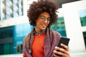 un hermoso pelo rizado chico de mezclado carrera es vestido en un de moda estilo juvenil bufanda y suéter escribe un mensaje en un telefono en contra el antecedentes de oficina edificios foto