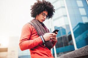 un hermoso pelo rizado chico de mezclado carrera es vestido en un de moda estilo juvenil bufanda y suéter escribe un mensaje en un telefono en contra el antecedentes de oficina edificios foto