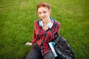 A beautiful red-haired student with freckles is dressed in a red checkered shirt with headphones sitting on the lawn in between the cheba. Student leisure photo