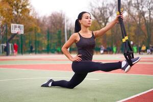 niña atleta formación utilizando trx en terreno de juego. mezclado carrera joven adulto mujer hacer rutina de ejercicio con suspensión sistema. sano estilo de vida. extensión al aire libre patio de juegos. foto