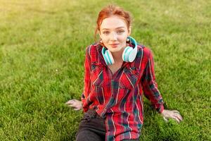 A beautiful red-haired student with freckles is dressed in a red checkered shirt with headphones sitting on the lawn in between the cheba. Student leisure photo