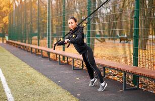 Girl athlete training using trx on sportground. Mixed race young adult woman do workout with suspension system. Healthy lifestyle. Stretching outdoors playground. photo