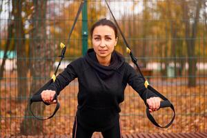 Girl athlete functional training on sportground. Mixed race young adult woman do workout with suspension system. Healthy lifestyle. Stretching outdoors playground. photo