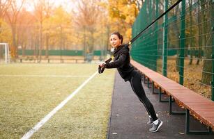 Girl athlete training using trx on sportground. Mixed race young adult woman do workout with suspension system. Healthy lifestyle. Stretching outdoors playground. photo