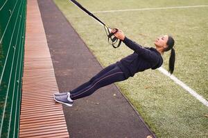 Girl athlete training using trx on sportground. Mixed race young adult woman do workout with suspension system. Healthy lifestyle. Stretching outdoors playground. photo