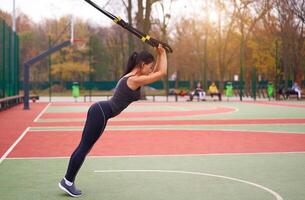 niña atleta formación utilizando trx en terreno de juego. mezclado carrera joven adulto mujer hacer rutina de ejercicio con suspensión sistema. sano estilo de vida. extensión al aire libre patio de juegos. foto