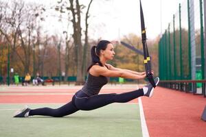 Girl athlete training using trx on sportground. Mixed race young adult woman do workout with suspension system. Healthy lifestyle. Stretching outdoors playground. photo
