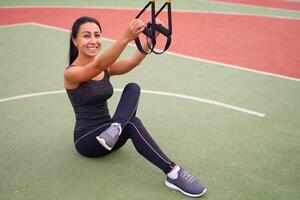 Girl athlete functional training on sportground. Mixed race young adult woman do workout with suspension system. Healthy lifestyle. Stretching outdoors playground. photo