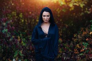A girl in a black dress, a cloak with a hood. It stands against the background of the autumn forest. Witch Costume, Satanist, Necromancer, Halloween Costume. photo