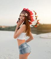 un hermosa joven caucásico niña en un blanco parte superior y mezclilla bermudas. en su cabeza vistiendo un indio sombrero cucaracha. en pie en el desierto. foto