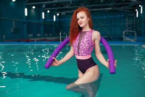 Beautiful young red-haired girl in a fashionable swimsuit in the style of the 80s stands with the noodles in her hands in the indoor swimming pool photo