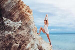 un hermosa joven niña con corto pelo es vestido en pantalones cortos y un blanco jersey practicando yoga en contra el fondo de el mar y montañas. actitud guerrero héroe. concepto acerca de equilibrar y calma foto