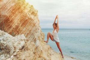 un hermosa joven niña con corto pelo es vestido en pantalones cortos y un blanco jersey practicando yoga en contra el fondo de el mar y montañas. actitud guerrero héroe. concepto acerca de equilibrar y calma foto