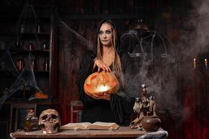 Halloween concept. Black witch holding Halloween pumpkin with carved smily face in hand standing dark room photo