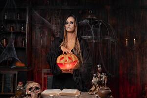 Halloween concept. Black witch holding Halloween pumpkin with carved smily face in hand standing dark room photo