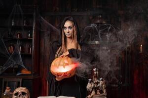 Halloween concept. Black witch holding Halloween pumpkin with carved smily face in hand standing dark room photo