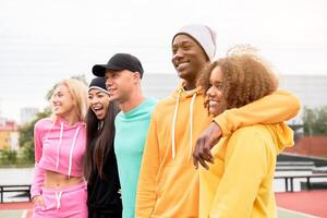 Group of multiethnic friends in sportsuits walking together at sport ground in city park, looking aside and laughing photo