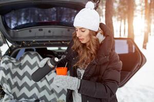 Young woman in woolen hat and black jacket stand neartrunk of the car and holds a cup of hot tea in her hands photo