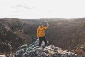 viajero en un amarillo chaqueta en pie en el borde de un acantilado tomando un selfie en su móvil teléfono a puesta de sol. divoká sarka valle, Praga foto