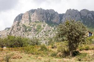 majestuoso rocoso montañas con lozano verde arboles y un distante de madera casa debajo un azul cielo foto