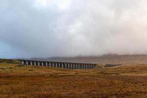 Misty landscape with an old viaduct crossing over a golden field under a cloudy sky at Ribblehead Viaduct, Yorkshire photo