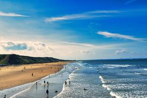 Sunny beach scene with people and rolling waves, blue sky with clouds overhead, and a hilly backdrop in Saltburn-by-the-Sea, England. photo