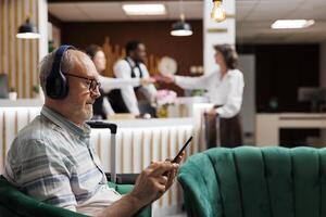 detallado ver de mayor masculino invitado relajante en acogedor salón zona con tableta y inalámbrico auriculares. retirado antiguo hombre escuchando a música y surf el red en digital dispositivo. de cerca, vista lateral. foto