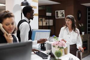 In hotel foyer, elderly guest arrives, checks-in, and receives assistance from friendly staff. Luggage is held by retired old woman while identification is checked by male receptionist at front desk. photo