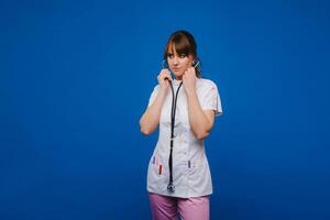 A female doctor, gesticulating, checks the heartbeat in the doctor's office at the hospital with a stethoscope isolated on a blue background photo