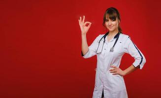 A young doctor in a white coat stands on a red background and shows with his hand that everything will be fine photo