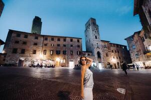 un niña en pantalones y un camiseta con lentes en el noche ciudad de san gimignano.a niña camina alrededor el ciudad en italia.toscana. foto