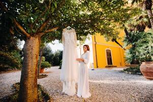 un hermosa novia soportes siguiente a un Boda vestir con un tocador atuendo siguiente a un villa en italia.mañana de el novia en toscana foto