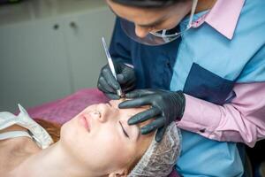 Procedure of eyebrow microblading. A master in black gloves is doing a blending needle of model's brow. photo