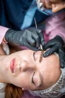 Procedure of eyebrow microblading. A master in black gloves is doing a blending needle of model's brow. photo