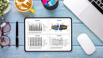 A business-focused desk with a tablet displaying graphs, a stylish cappuccino, laptop, and office supplies on a blue wooden background. photo