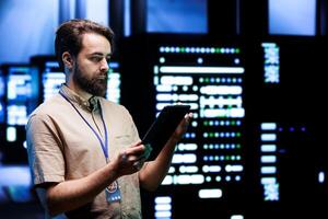 Worker doing system overhaul, imputing data on tablet of required patching in server cabinets. Facility housing advanced data storage equipment supporting critical IT workloads photo