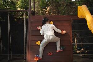 niño alpinismo en de madera pared a patio de juegos. joven niño ascendente un alpinismo pared con vistoso asideros a un al aire libre patio de juegos. actividad para niño patio de recreo concepto. foto