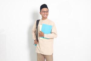 Portrait of excited student Asian muslim man in koko shirt with skullcap carrying backpack, while holding his school books. Islamic education concept. Isolated image on white background photo